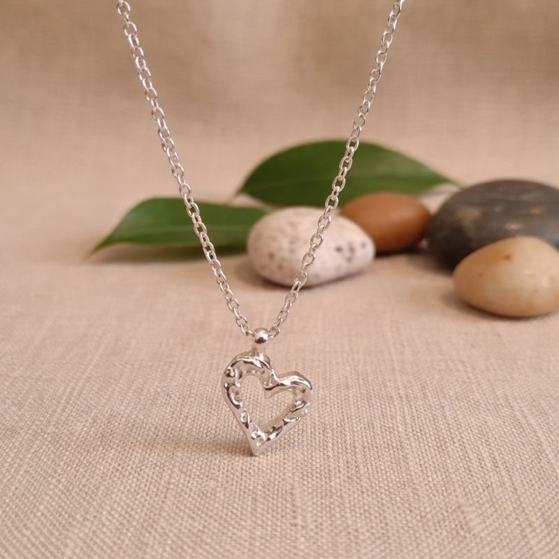 he Sculpted Heart Silver Necklace in 925 sterling silver, presented on a linen surface with natural river stones and green leaves.