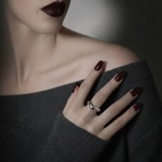Close-up portrait of a woman with dark burgundy lips and matching manicure, wearing the Liquid Drop silver ring. The polished 925 sterling silver ring reflects the light, creating a bold contrast against the dark tones of her outfit and makeup